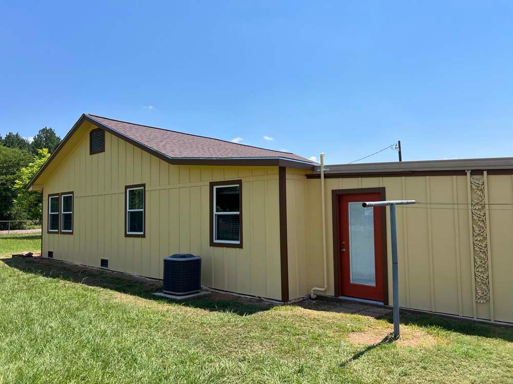 Small beige building with brown roof, red door, and windows beside a grassy field