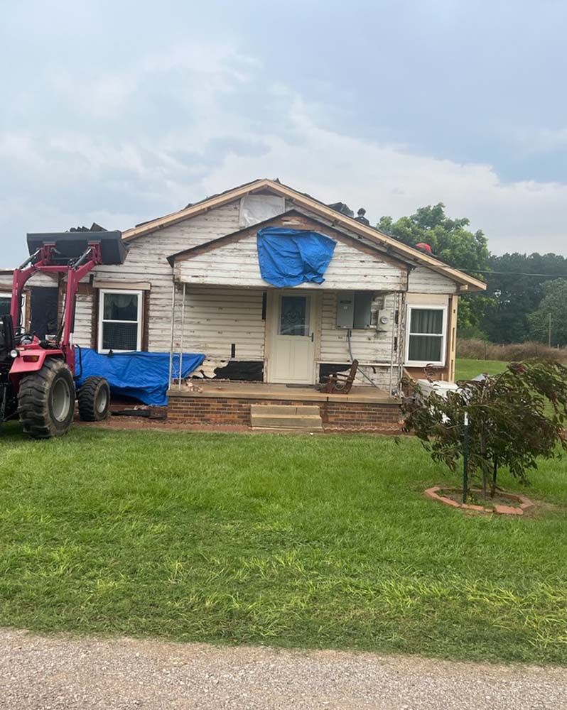 Small white house with blue tarp covering the roof, beside a red tractor on a grassy yard.
