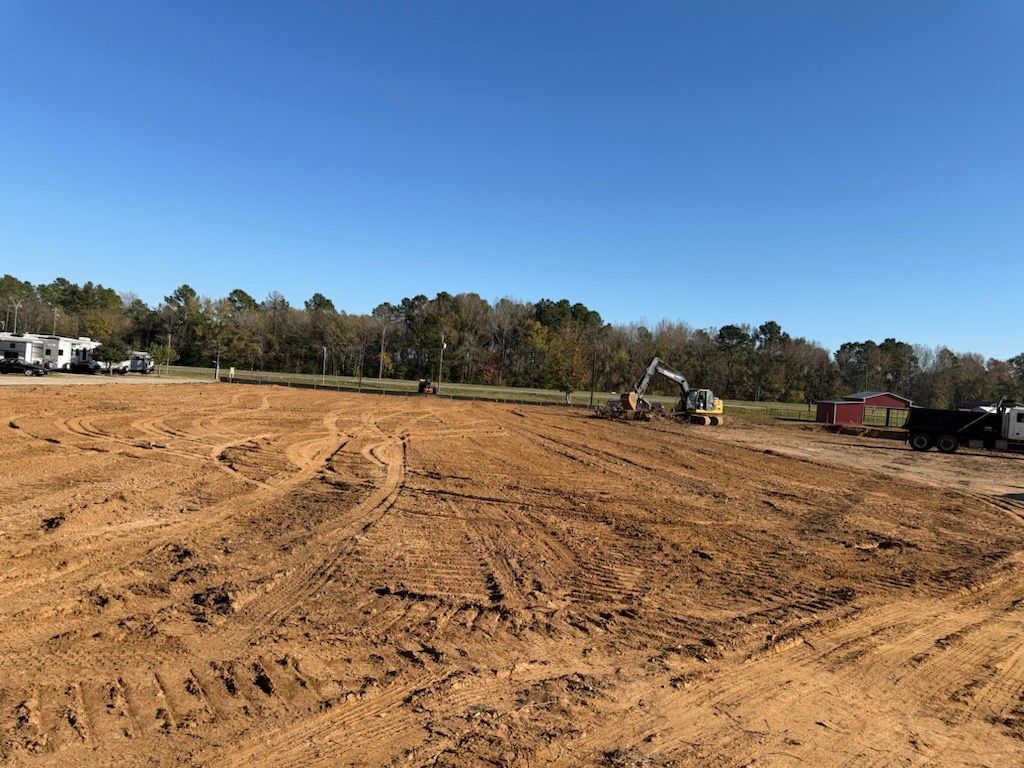 Wide dirt construction site with excavators, trucks, and a tree line under a clear blue sky