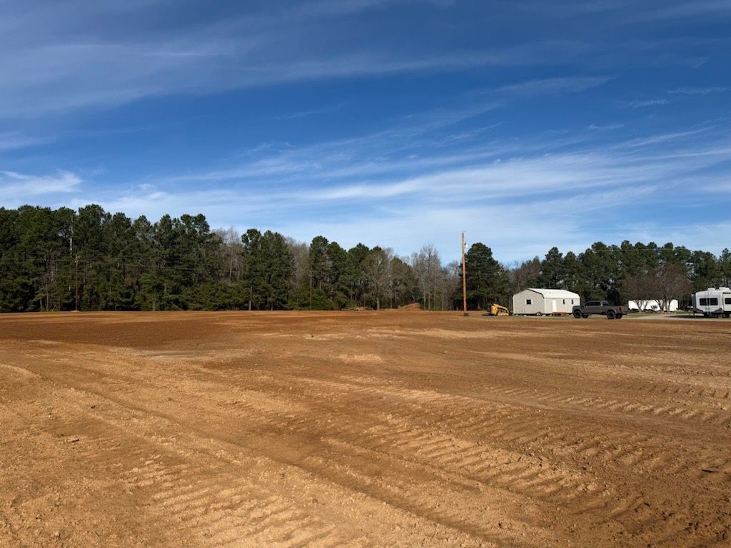 Open dirt lot with tire tracks, pine trees, and small buildings under a blue sky