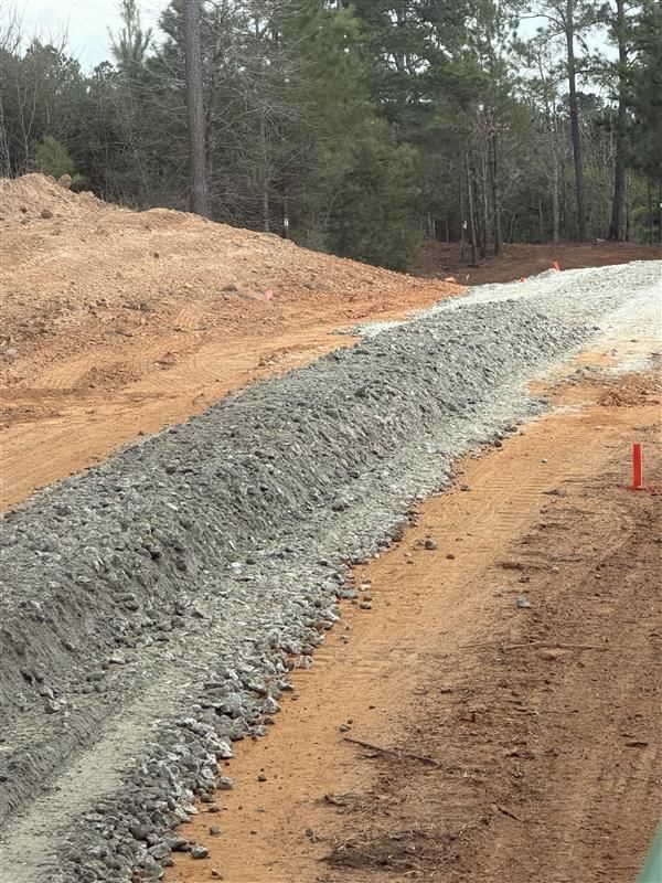 Gravel road under construction through a wooded area, with orange dirt and a red marker stake.