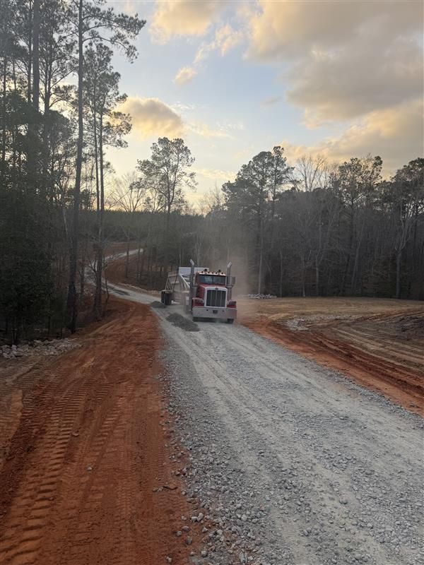 Gravel roadwork scene with a utility truck on a red dirt road at sunset, surrounded by pine trees.