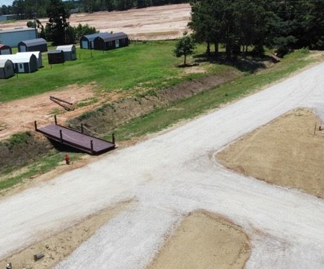 Gravel road junction beside grassy lots, small buildings, and trees near a rural construction area