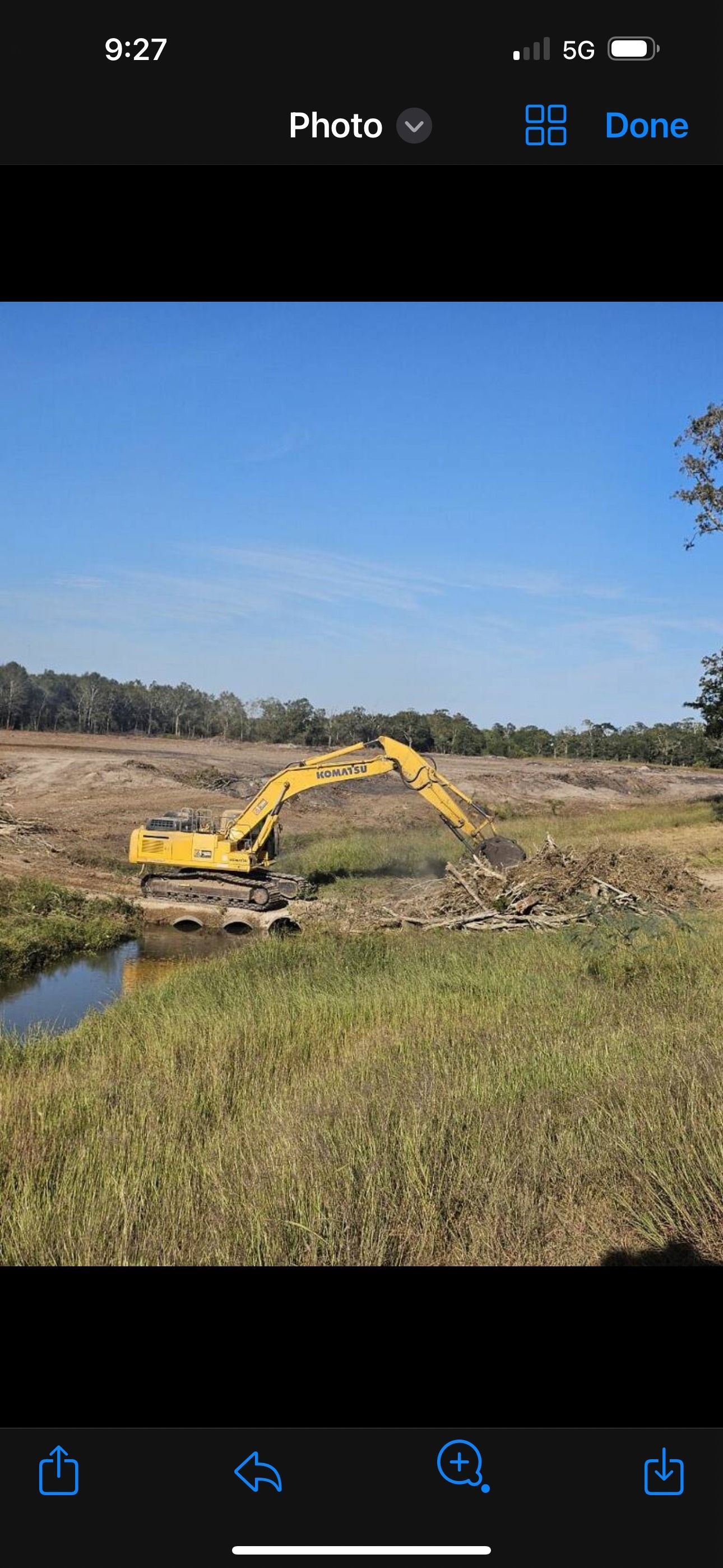 Yellow excavator working beside a narrow water channel in a grassy, open field under a clear blue sky