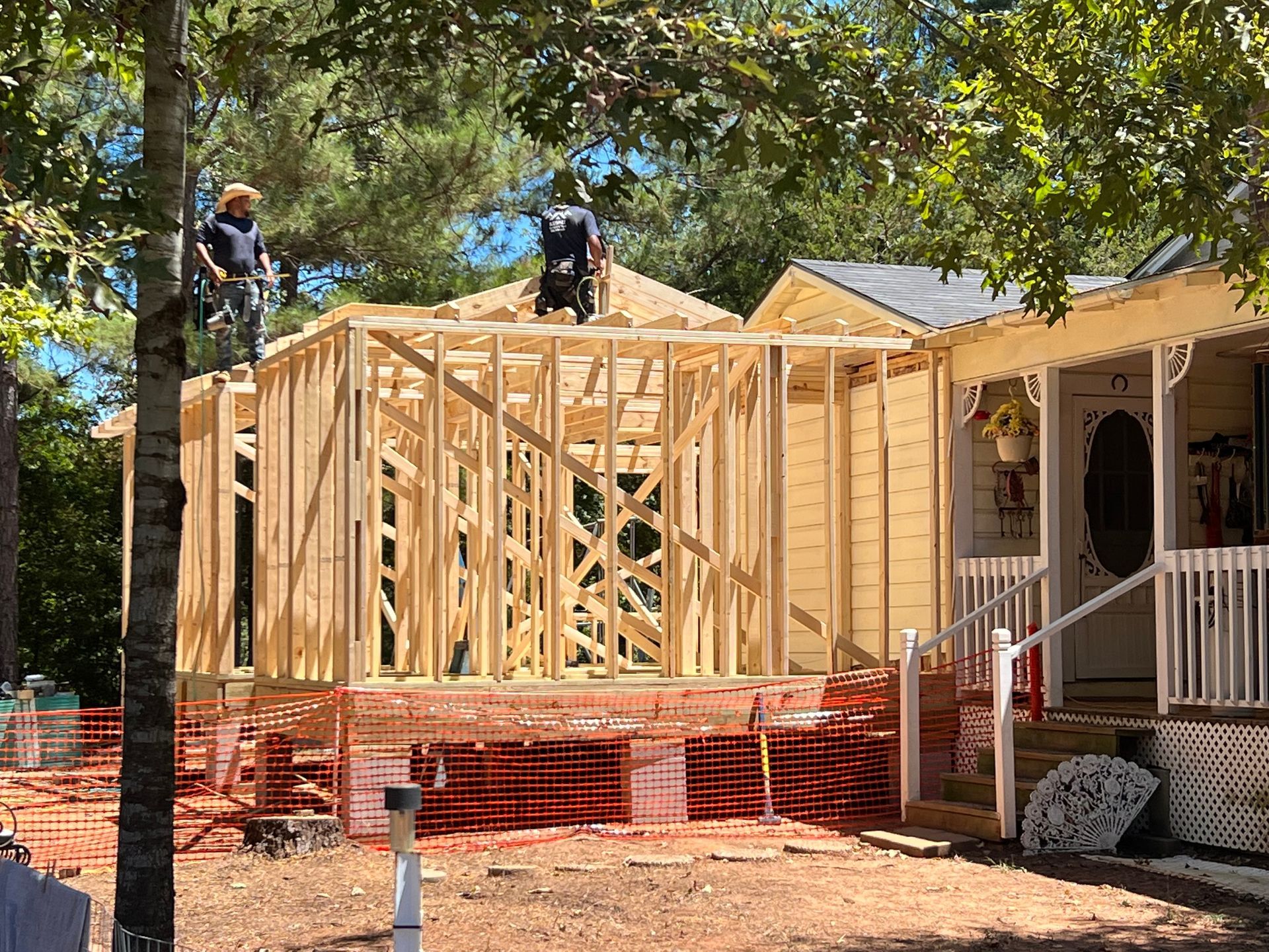 Two workers framing a house addition beside a porch, with orange safety fencing and trees overhead.
