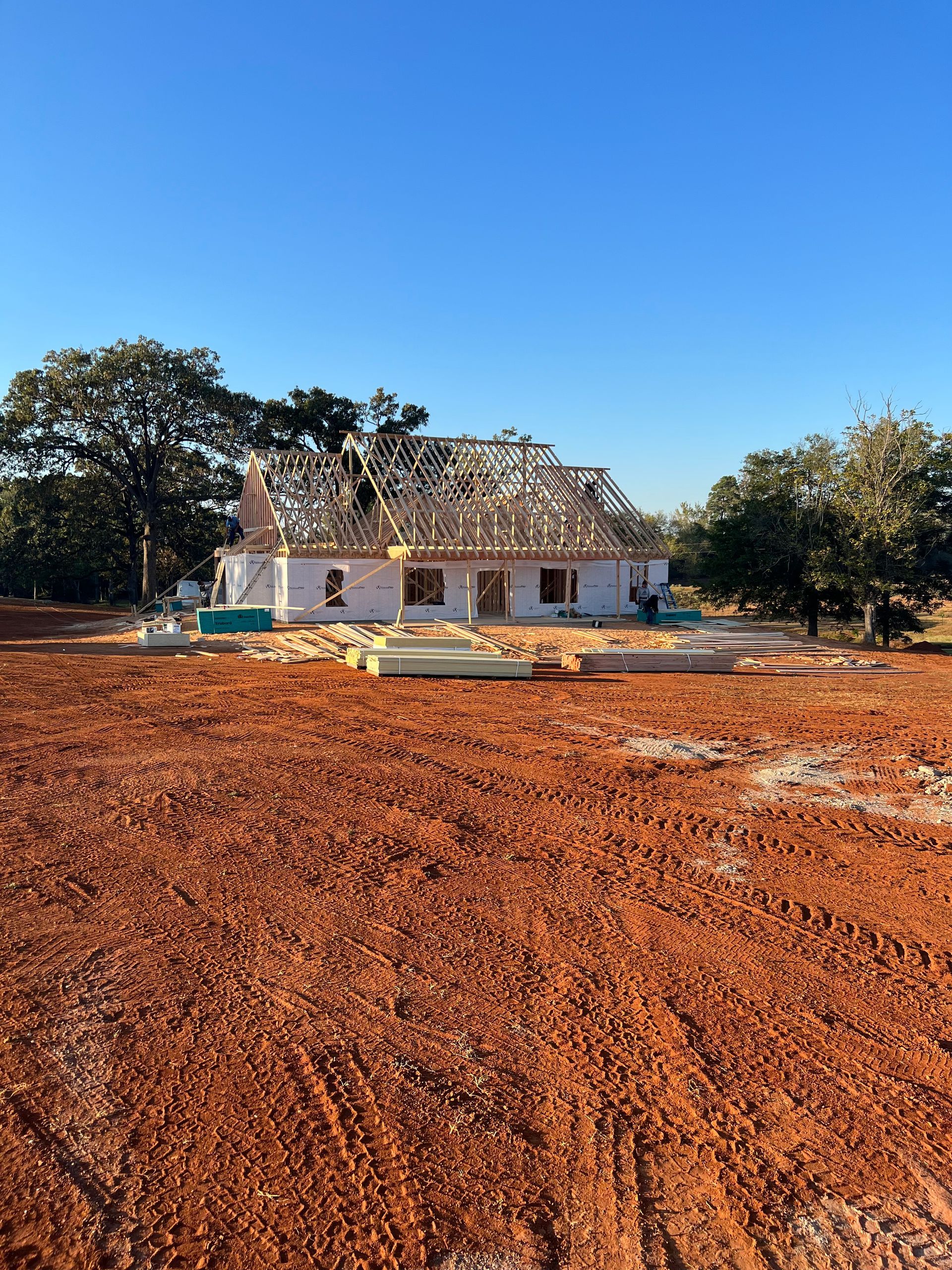Single-story white house under construction on red dirt, with trees and a clear blue sky.