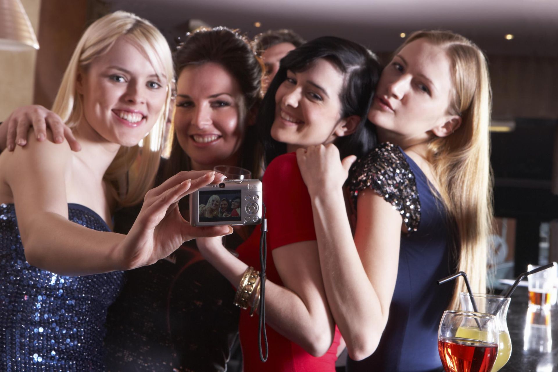 Four women smiling, taking a photo with a digital camera in a bar setting. Drinks on the bar are visible.