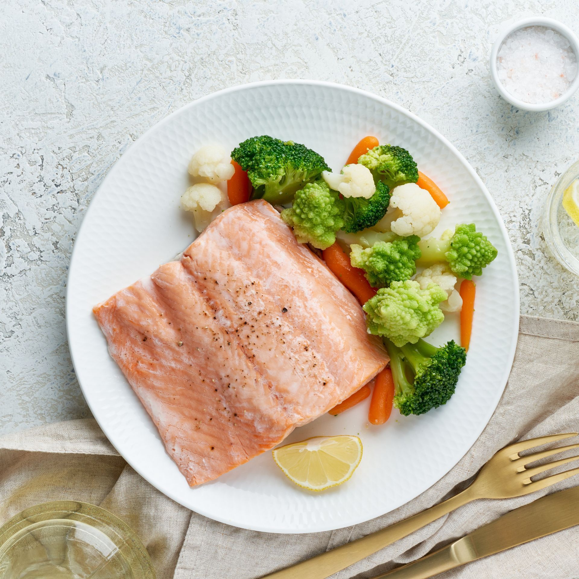 Um prato branco coberto com salmão, brócolis, cenoura e couve-flor. Sugestão de almoço saudável.