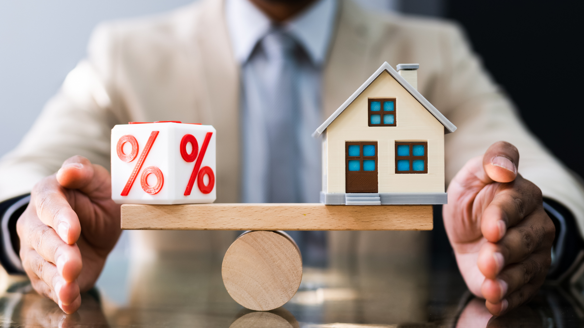 A man is holding a house and a percentage cube on a wooden balance beam.