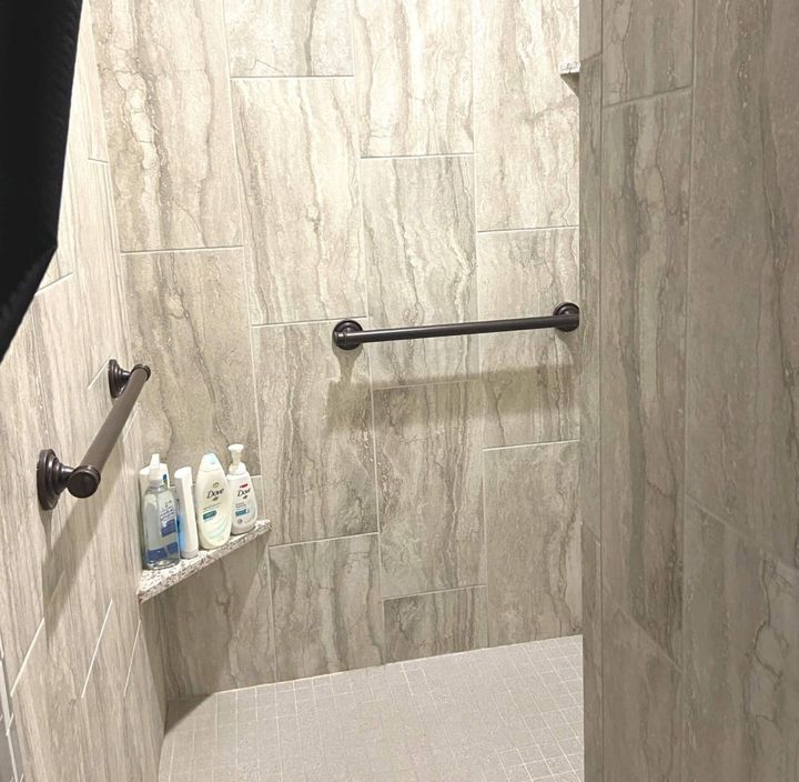 Shower with gray tile walls, grab bars, and built-in shelf with toiletries.