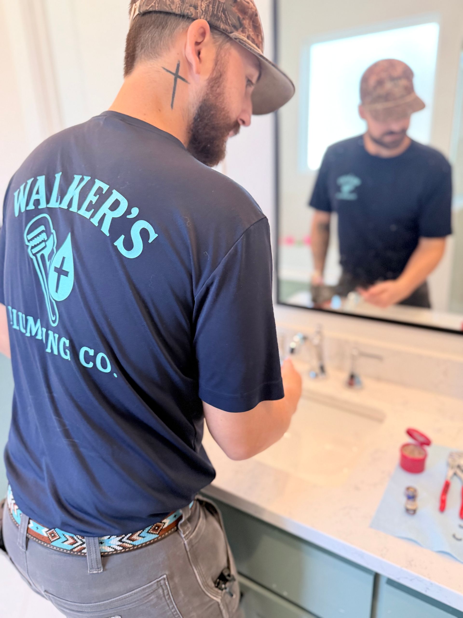 Man in navy shirt and cap washing hands at a bathroom sink, seen from behind and reflected in the mirror