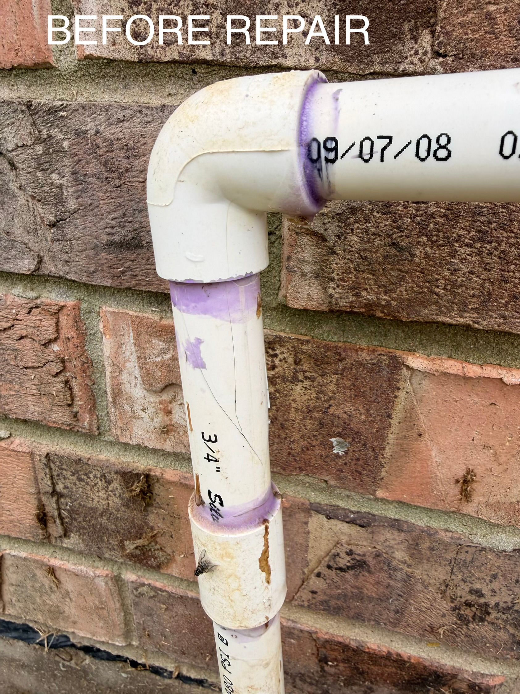 White PVC pipe elbow joint on a brick wall, labeled “before repair,” with purple primer marks.