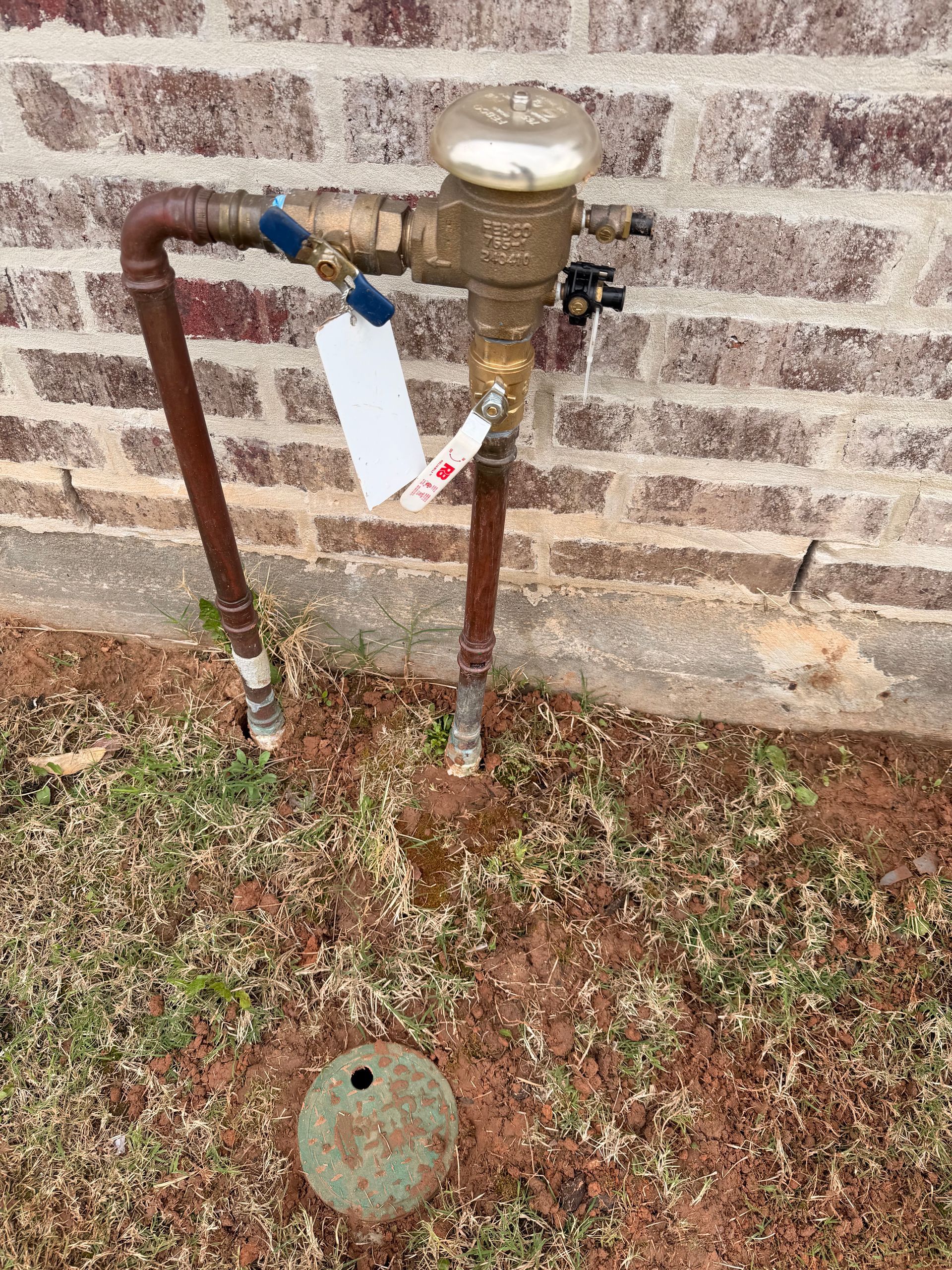 Outdoor water meter and rusty pipes against a brick wall, with a valve handle and hanging tag.