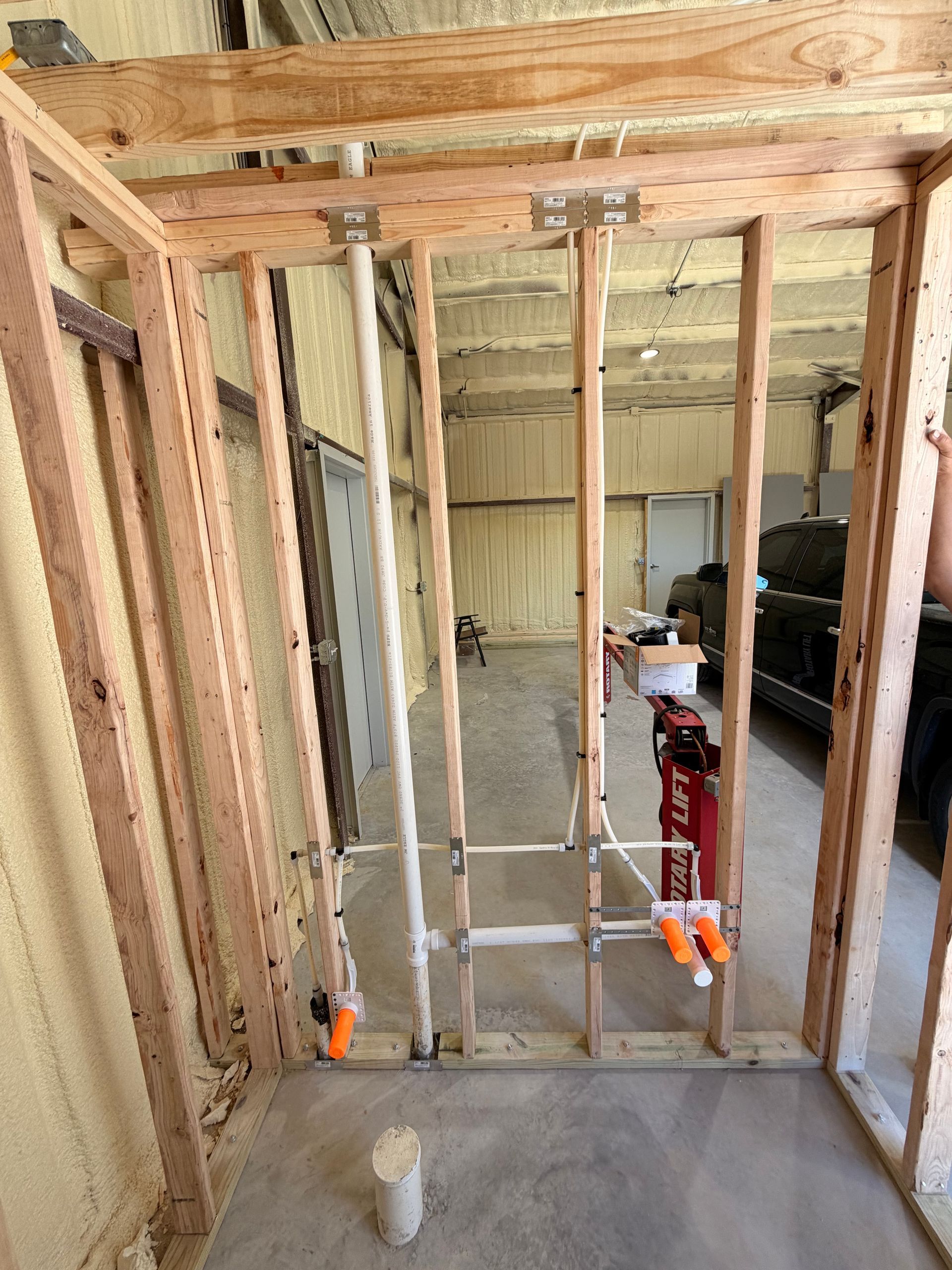 Framed interior wall under construction with exposed studs, pipes, and clamps in a garage-like room