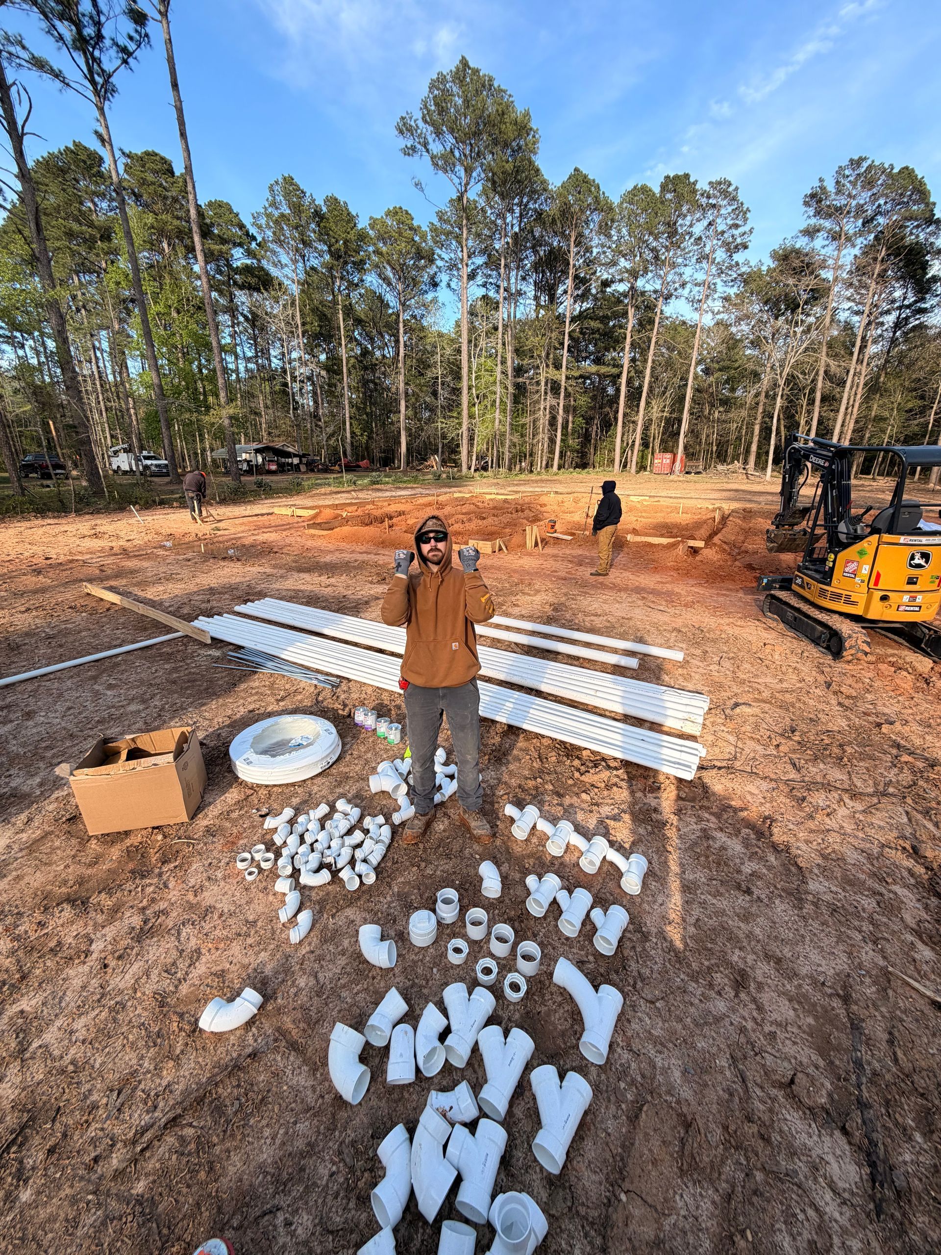 Person standing beside white panels and parts laid out on gravel in a wooded construction site.