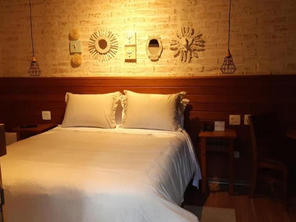 Quarto com iluminação aconchegante, cama branca impecavelmente arrumada, cabeceira de madeira e obras de arte na parede acima dela.