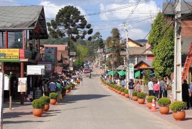 Rua de pedestres com lojas, vasos de plantas e pessoas caminhando em um dia ensolarado.