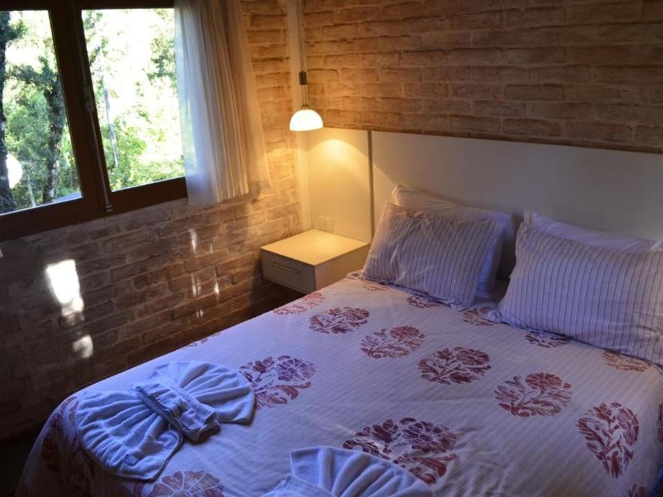 Quarto aconchegante com parede de tijolos, luz natural na janela, cama, abajur e toalhas dobradas sobre a cama.