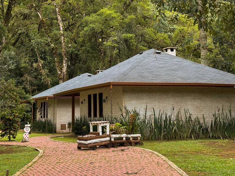 Pequena casa bege com telhado cinza ao lado de um caminho de tijolos em um parque arborizado.