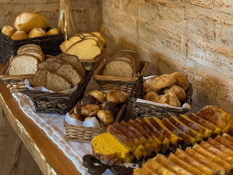 Cestas com pães e doces variados sobre uma mesa encostada em uma parede de tijolos.