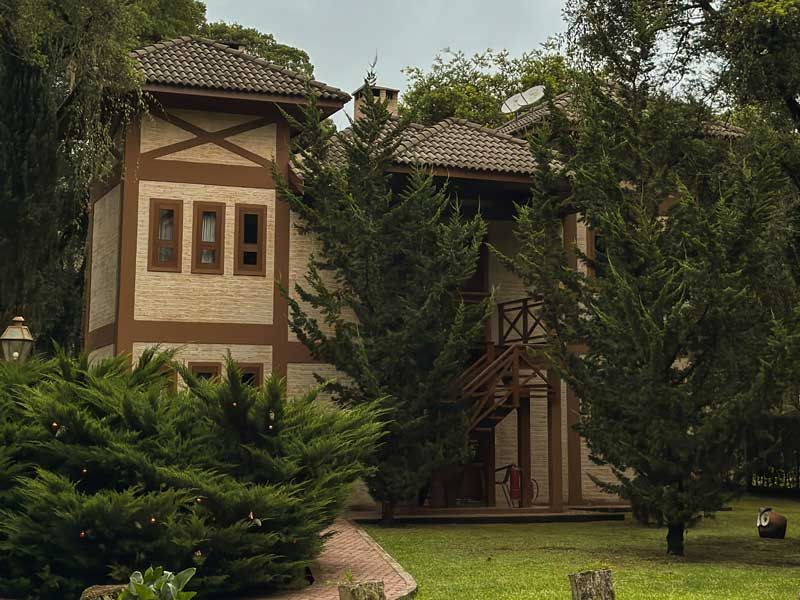 Casa de dois andares com telhado de telha e vegetação exuberante, com árvores e arbustos em primeiro plano.