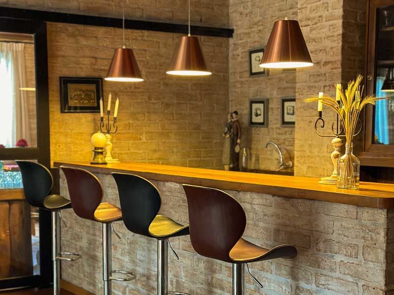 Bar aconchegante com paredes de tijolos aparentes, balcão de madeira, banquetas pretas e luminárias pendentes de cobre.