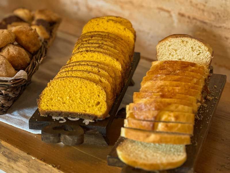 Fatias de pão amarelo e branco dispostas em uma bandeja ao lado de pãezinhos à esquerda.
