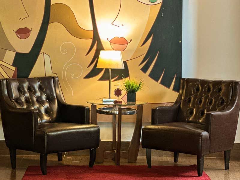 Duas cadeiras marrom-escuras ao lado de uma pequena mesa com um abajur, encostadas em uma parede com um mural estilizado.