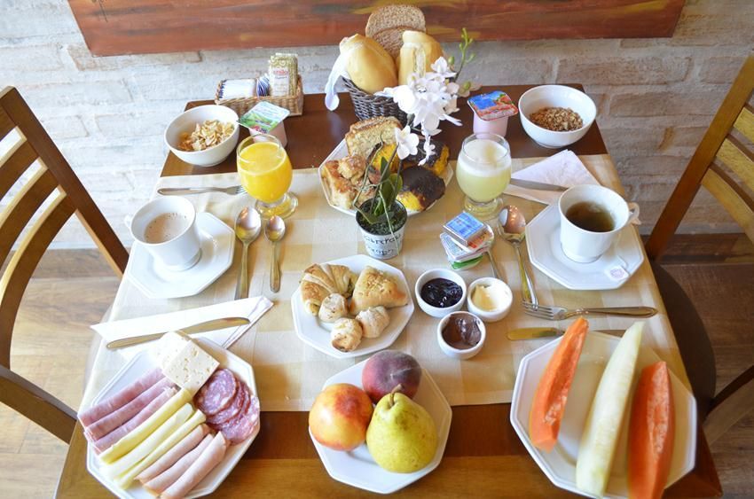 Mesa de café da manhã com diversos doces, frutas, sucos, café e flores sobre uma toalha de mesa branca.