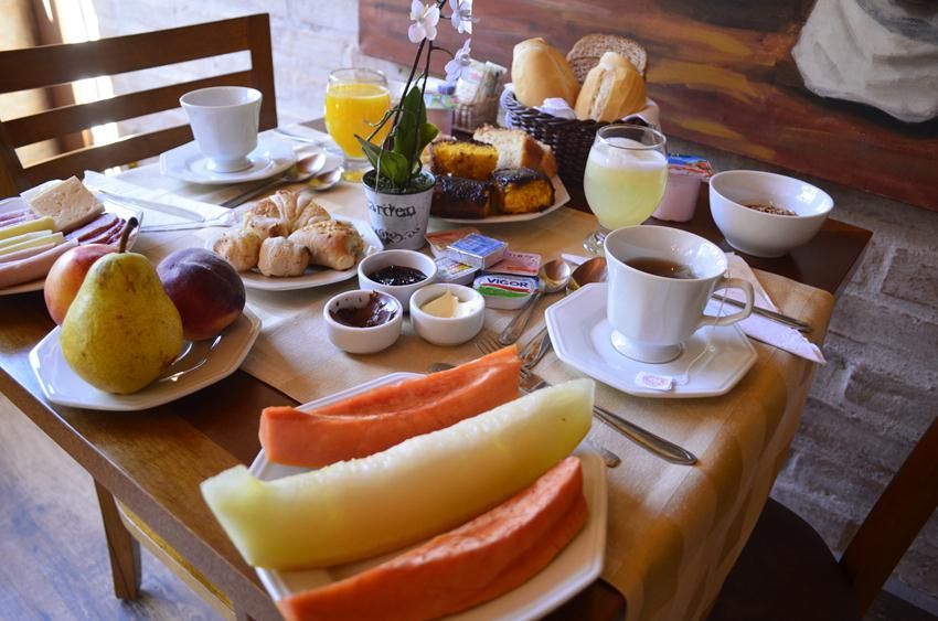 Café da manhã servido em uma mesa de madeira com frutas, doces, café e suco em uma sala de jantar aconchegante.