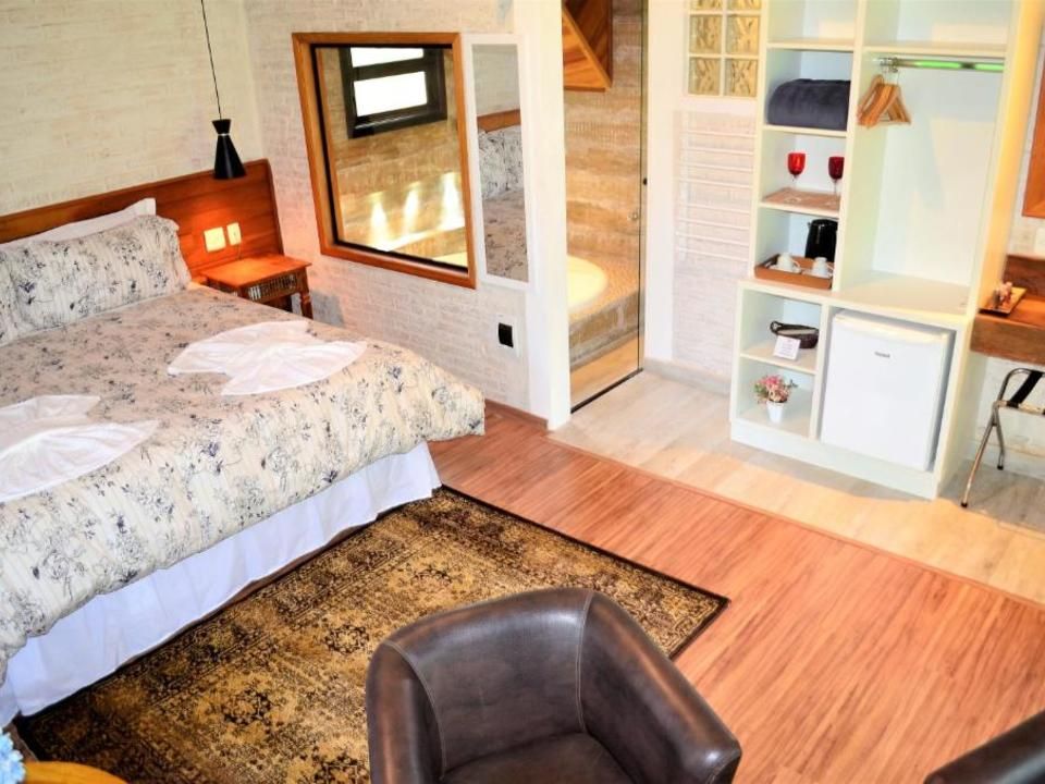 Quarto aconchegante com cama florida, cadeira marrom, tapete e prateleiras abertas junto a uma janela ensolarada.