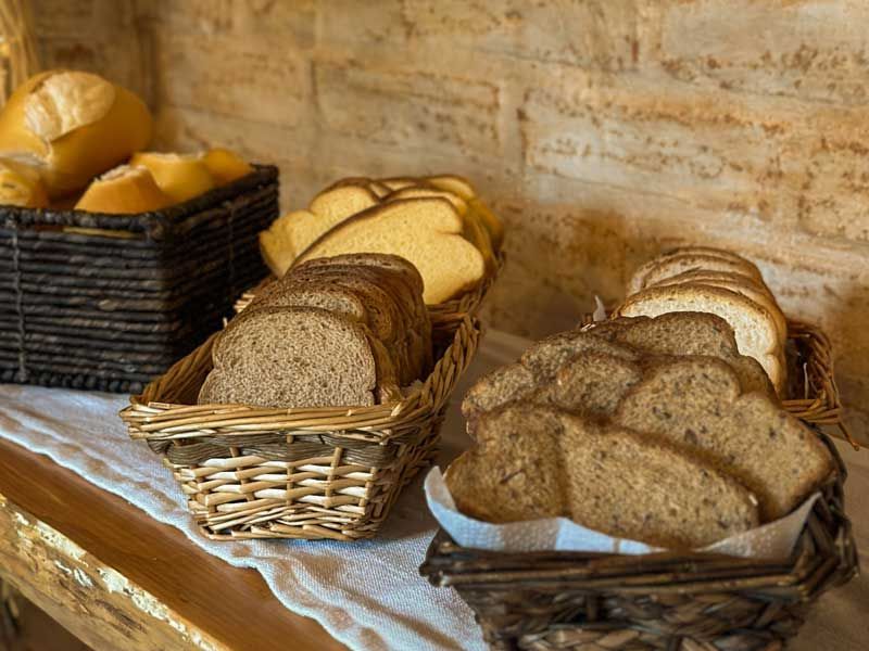 Cestas de pães fatiados variados sobre uma mesa de madeira encostada em uma parede rústica.