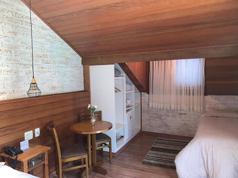 Quarto aconchegante no sótão com teto de madeira, cama branca, escrivaninha, prateleiras e tapete listrado.