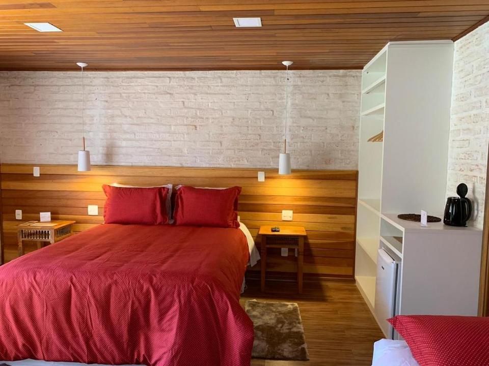 Quarto com colcha vermelha, teto de madeira, parede de tijolos brancos e estante ao lado da cama.
