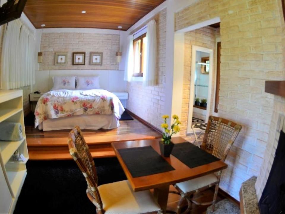 Quarto aconchegante com paredes de tijolos brancos, teto de madeira, cama e uma pequena mesa com cadeiras em frente a um espelho.