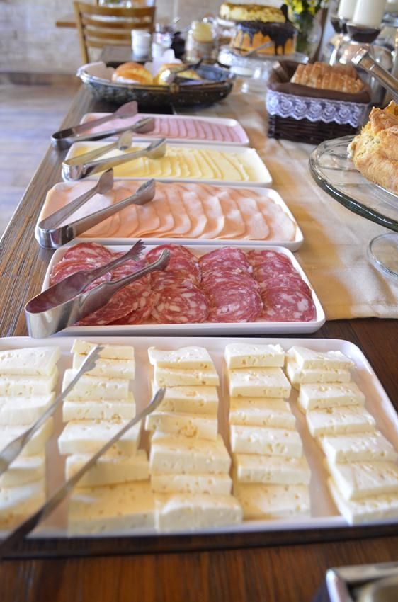Mesa de bufê com uma variedade de queijos fatiados e frios em bandejas brancas, servidos com pinças.