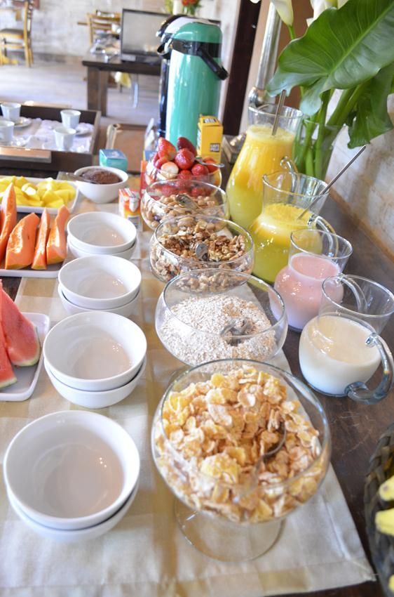 Buffet de café da manhã com frutas, iogurte, doces e uma garrafa térmica azul-turquesa sobre a mesa.
