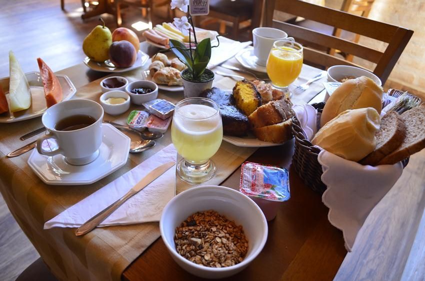 Mesa de café da manhã com café, suco, pão, frutas, iogurte e granola em uma sala de jantar ensolarada.