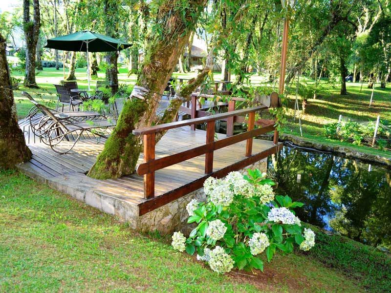 Caminho de jardim com uma pequena ponte de madeira sobre um riacho, hortênsias e mesas sob um guarda-sol verde.