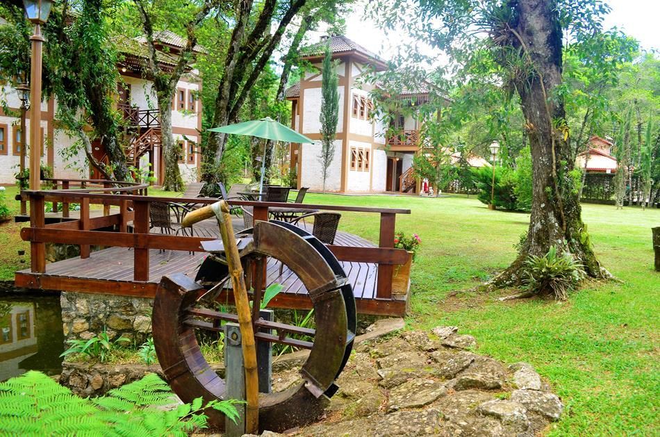 Roda d'água rústica ao lado de uma ponte de madeira em um jardim exuberante com chalés e árvores.