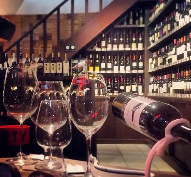 Taças e garrafas de vinho sobre uma mesa de restaurante, com uma prateleira de vinhos ao fundo.