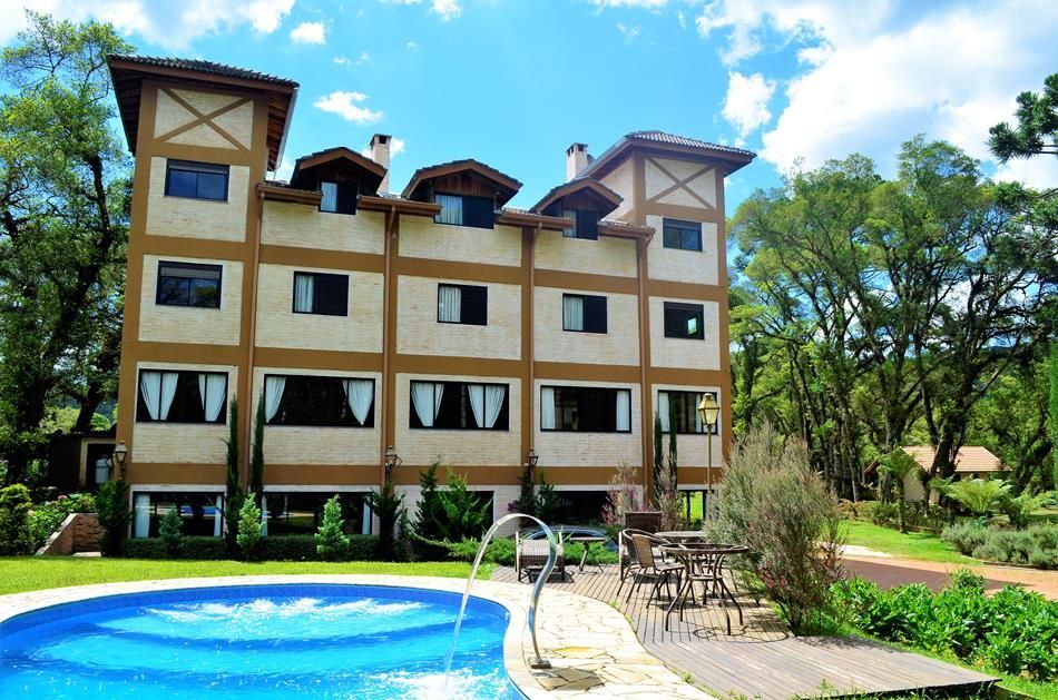 Edifício de resort bege com varandas ao lado de uma piscina azul e pátio, rodeado por árvores verdes.