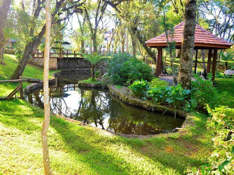 Jardim exuberante com um pequeno lago, ponte de madeira e gazebo de telhado vermelho entre as árvores.