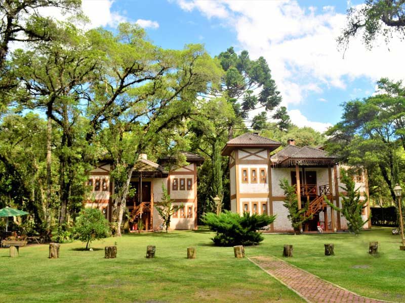 Casa histórica entre grandes árvores verdes em um gramado ensolarado sob um céu parcialmente nublado.