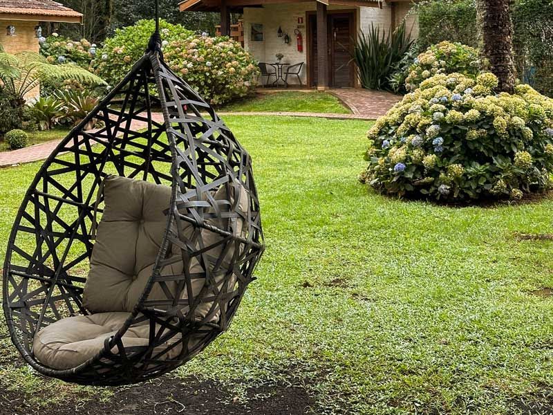 Cadeira suspensa de vime preto com almofada sobre um gramado verde em um jardim.