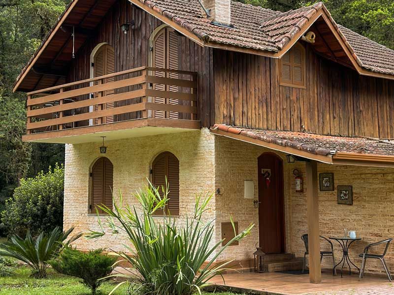 Chalé rústico de madeira de dois andares com piso térreo de pedra, varanda e alpendre em meio a uma vegetação exuberante.