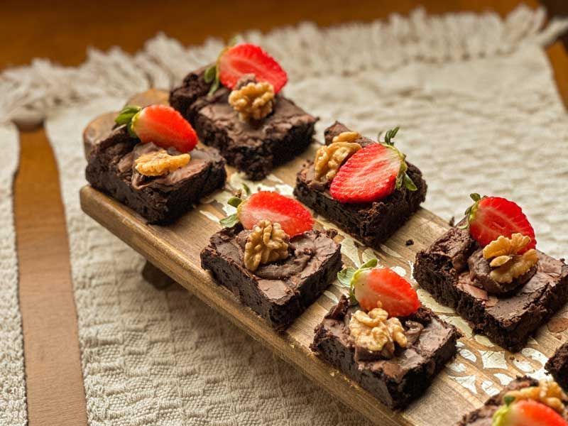 Quadrados de sobremesa de chocolate cobertos com morangos, dispostos em uma tábua de madeira e servidos sobre um pano texturizado.