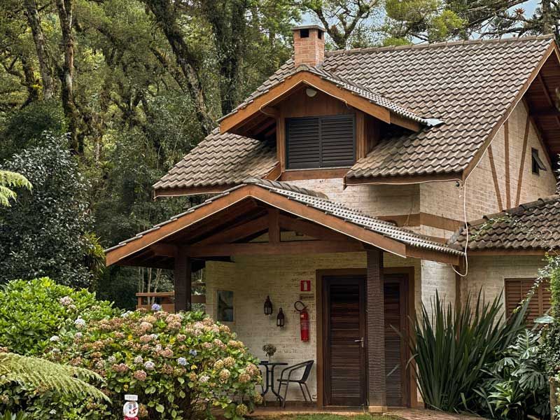 Chalé aconchegante com telhado de telha, varanda de madeira e um jardim exuberante com árvores e arbustos.