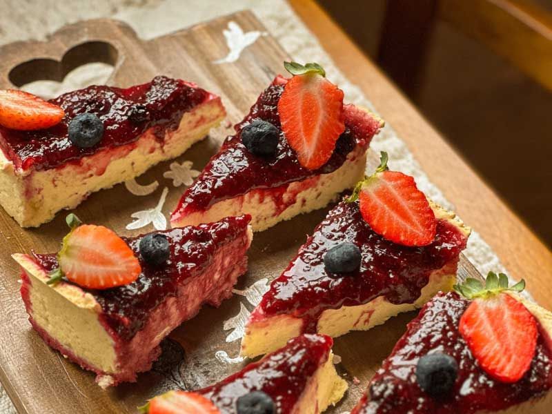 Fatias de cheesecake com cobertura de frutas vermelhas servidas em uma tábua de madeira, decoradas com morangos e mirtilos.