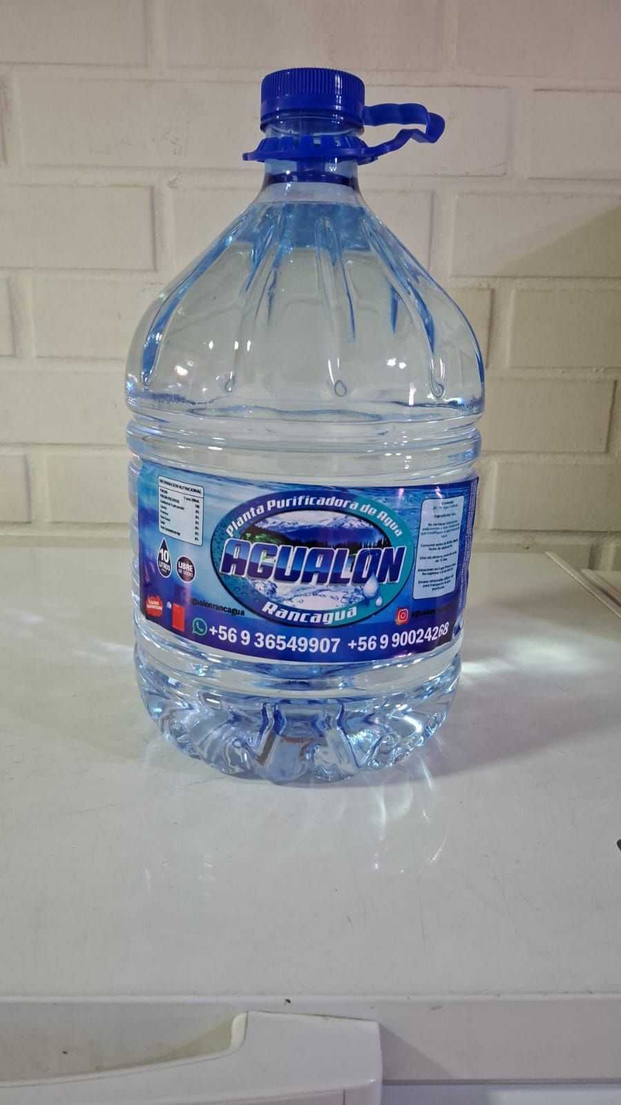 Jarra de agua grande de plástico transparente con etiqueta y tapón azules sobre una mesa blanca contra una pared de azulejos.